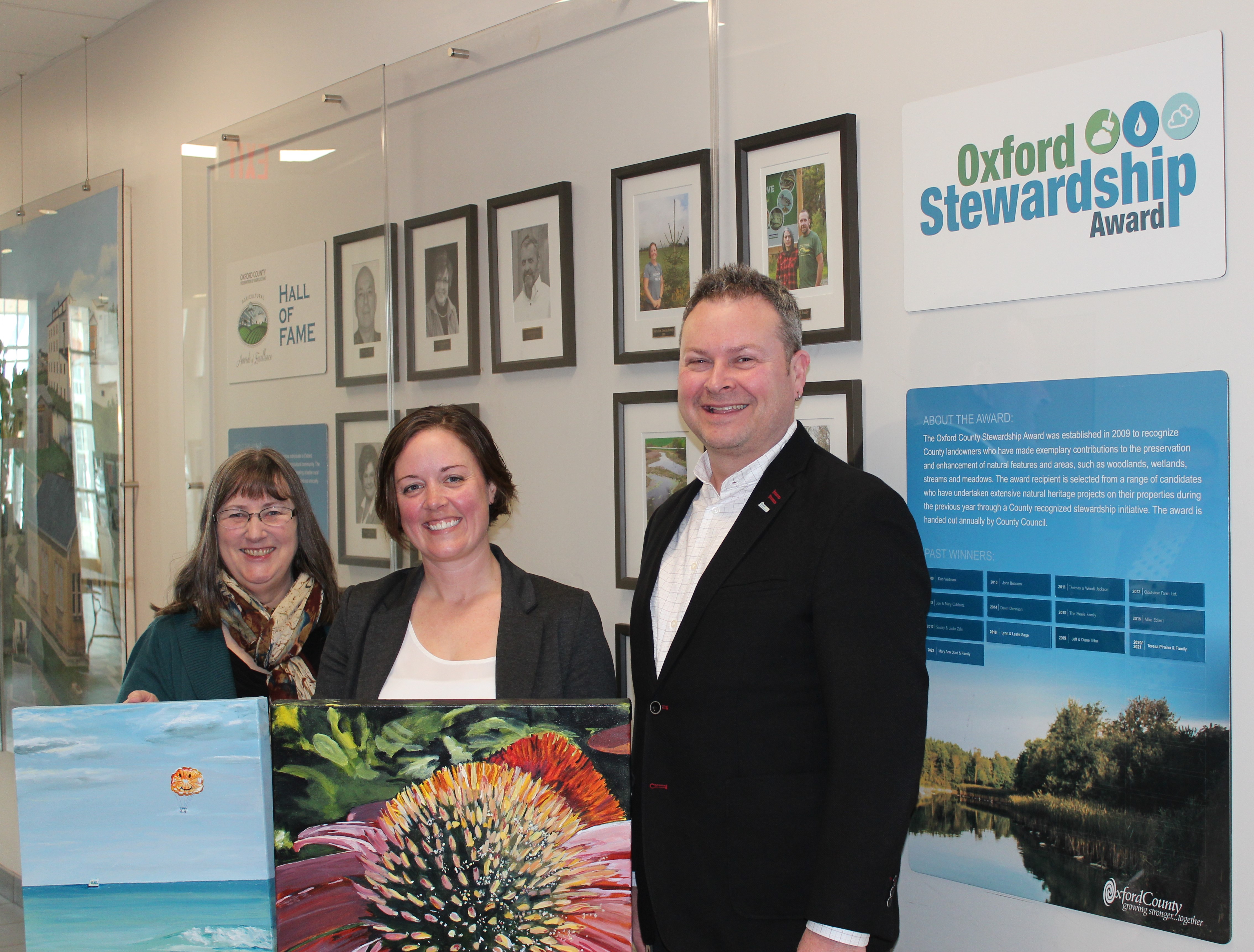 Vonnie Snyder Mary Anne Dore and Warden Ryan with original artwork in front of the Oxford Stewardship Award wall 