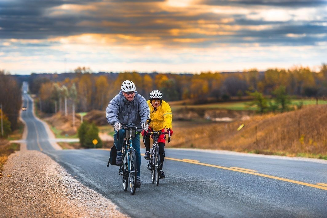 2 men on bicycles riding on a paved road in fall with fields on either side