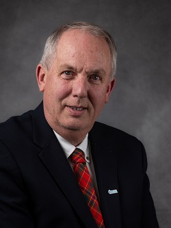 Councilor David Mayberry, wearing a dark blue suit with a red, green and yellow plaid tye, he has short white and brown hair. 
